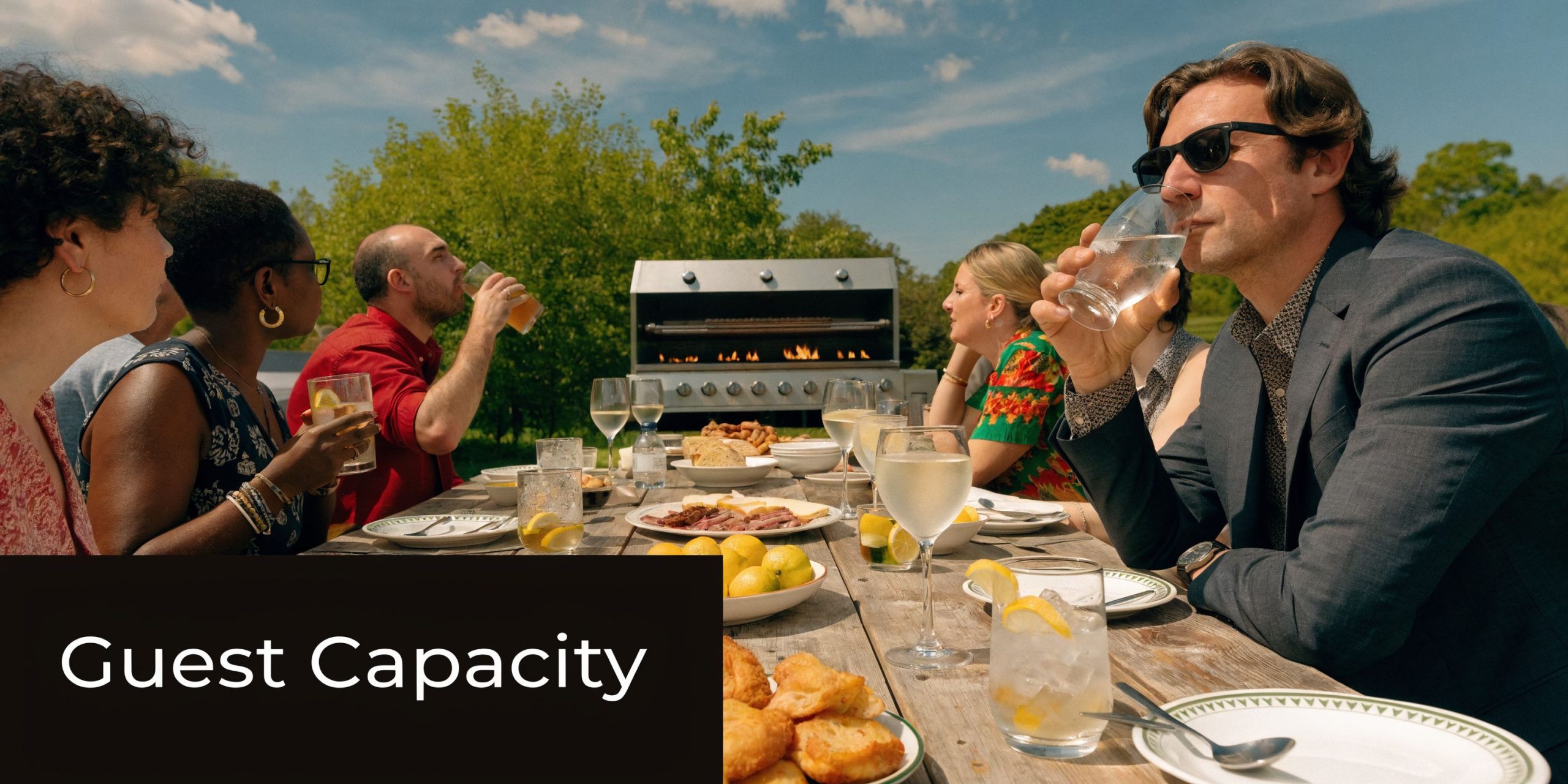 A group of friends enjoy an outdoor garden barbecue dinner around a wooden table in the sunshine.