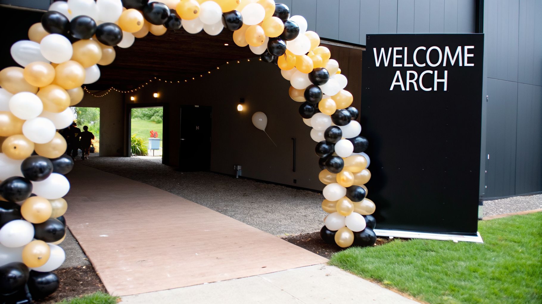 A black, white, and gold balloon arch welcomes guests into a building entrance with a sign.