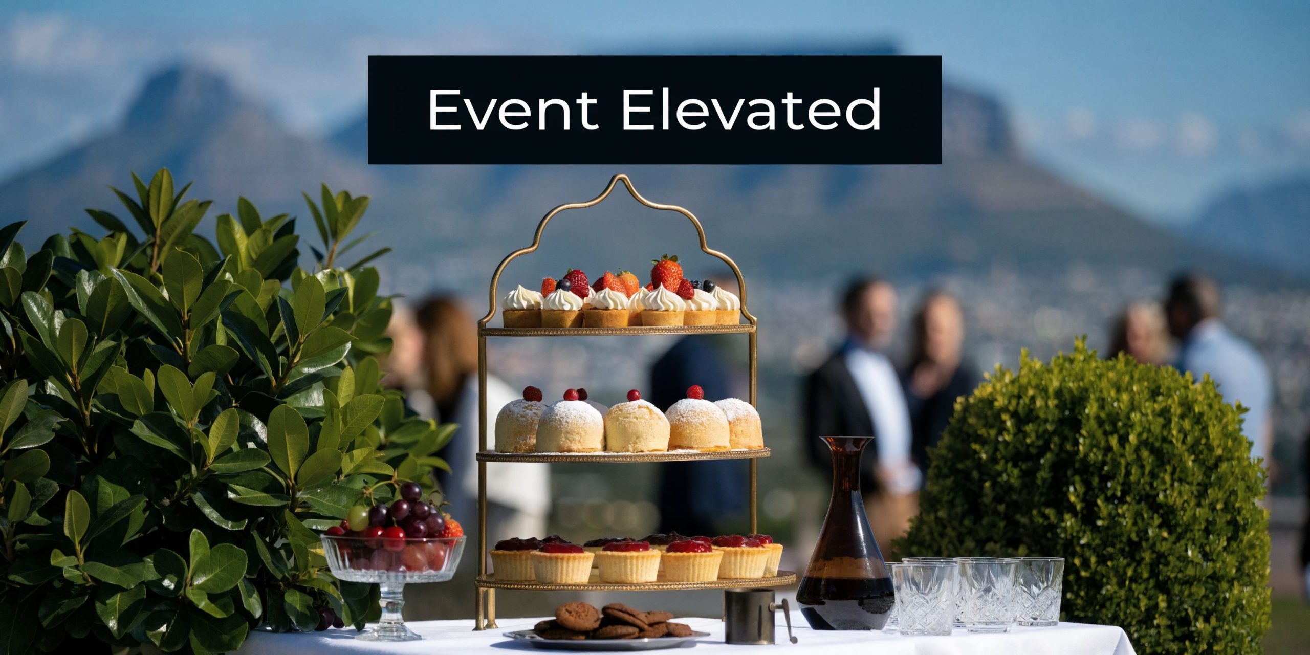 A three tier cake stand filled with assorted gourmet desserts set against a scenic mountain landscape backdrop.