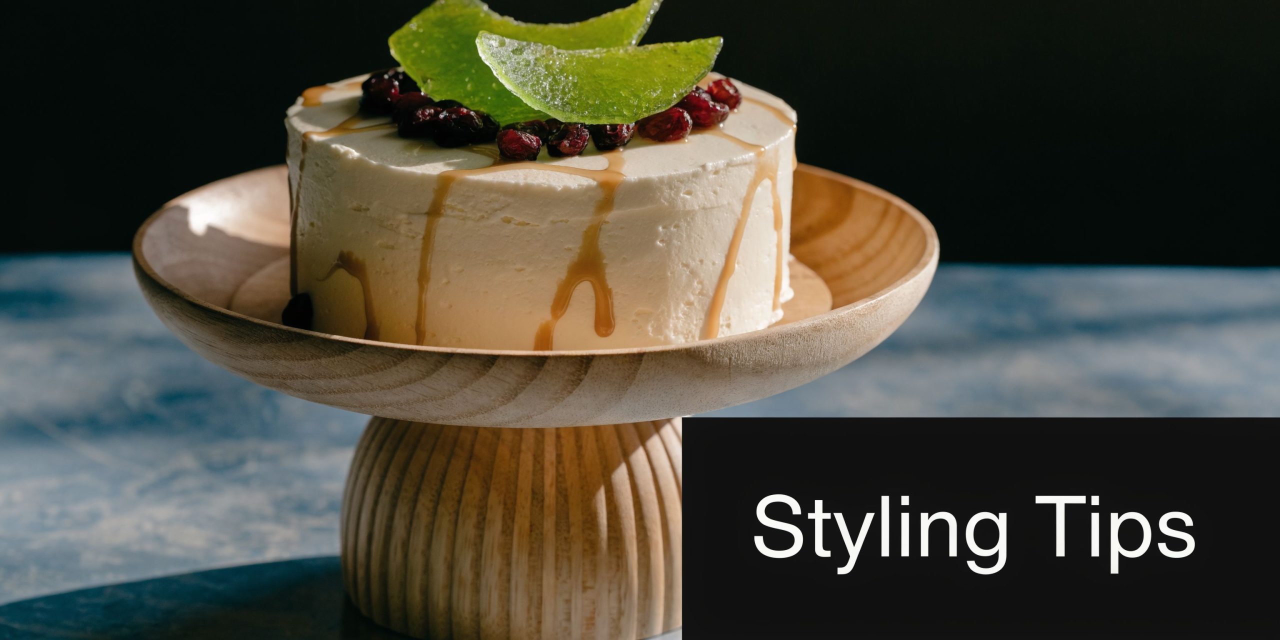 A creamy cake topped with dried fruit on a stylish wooden cake stand against a dark background.