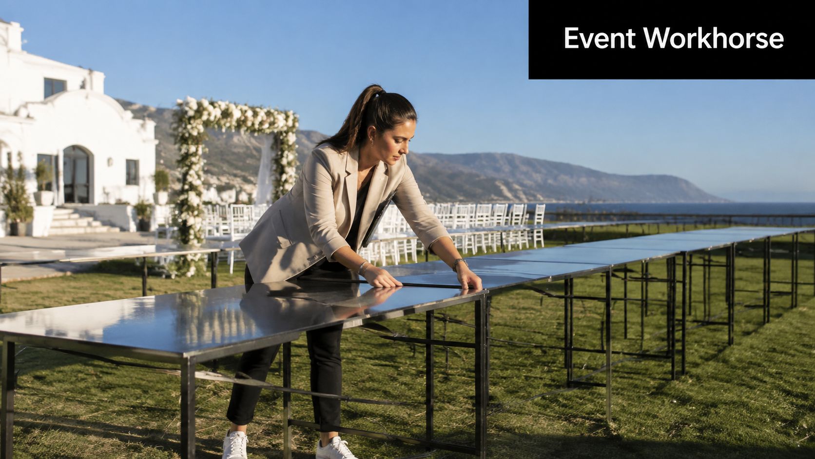 A woman carefully assembling a long, reflective steel trestle table at an outdoor wedding event venue.